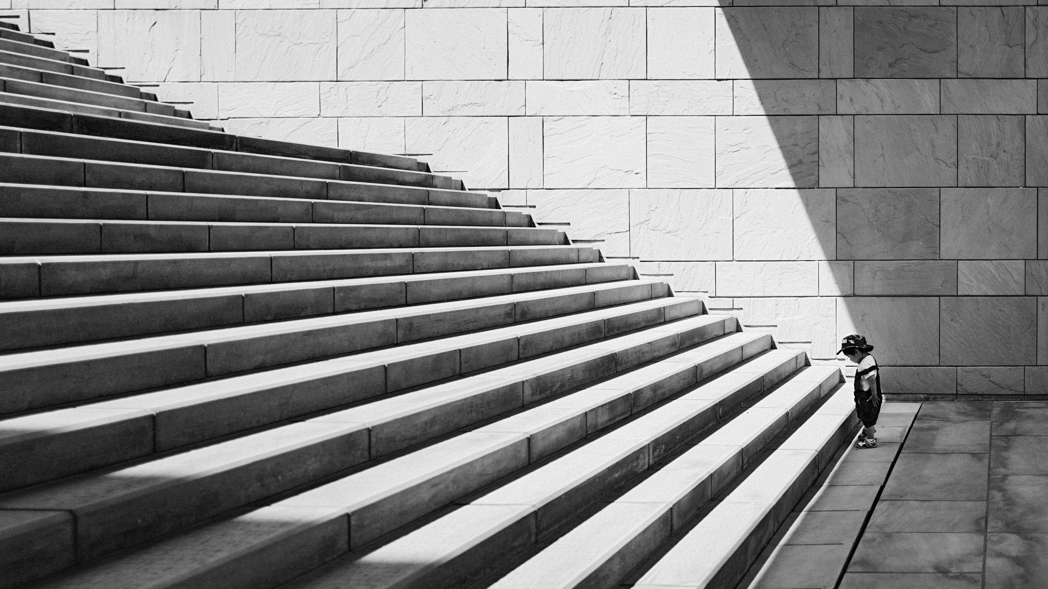 A small child standing at the bottom of a large set of concrete stairs in black and white